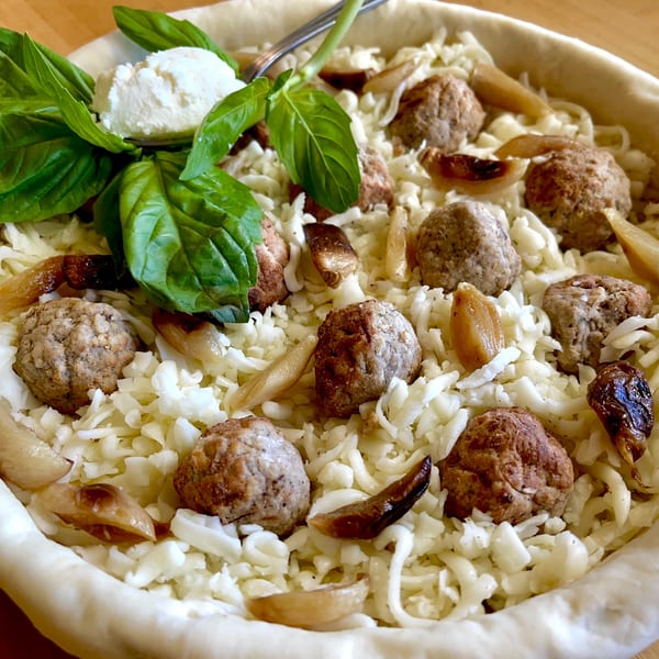 Unbaked Italian deep dish pizza with meatballs, cheese, roasted garlic, and fresh basil.