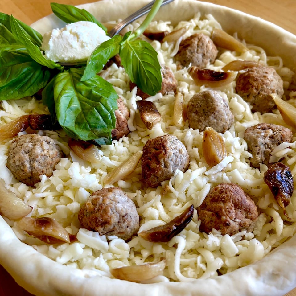 Unbaked Italian deep dish pizza with meatballs, cheese, roasted garlic, and fresh basil.
