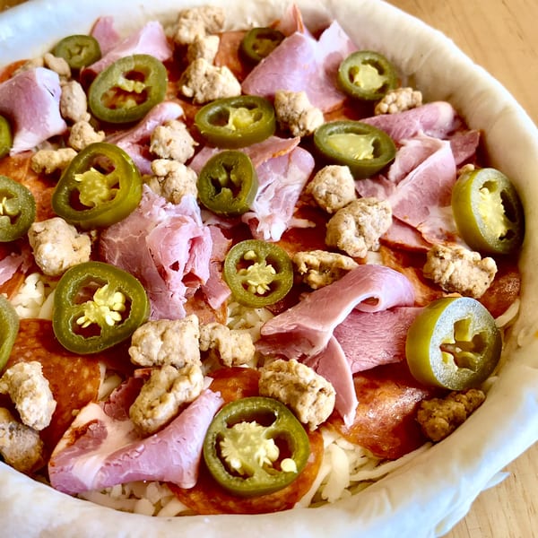 Unbaked deep-dish pizza with pepperoni, ham, sausage, jalapeños, and cheese in a white crust.