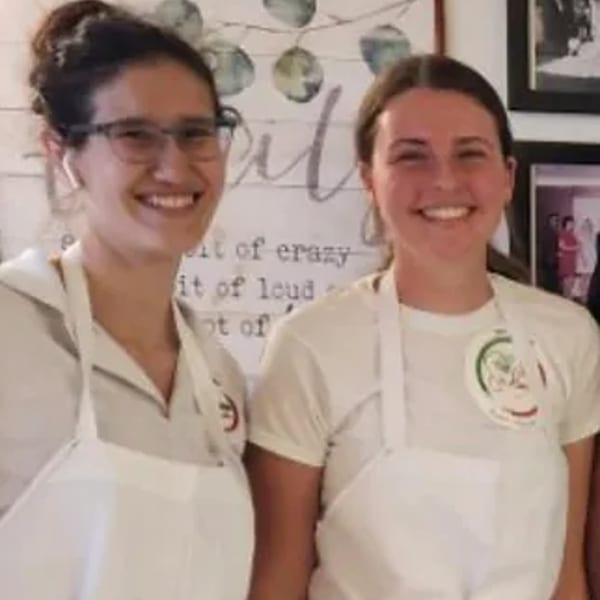 Two smiling women in white aprons, one wearing glasses, standing in front of a decorative wall.