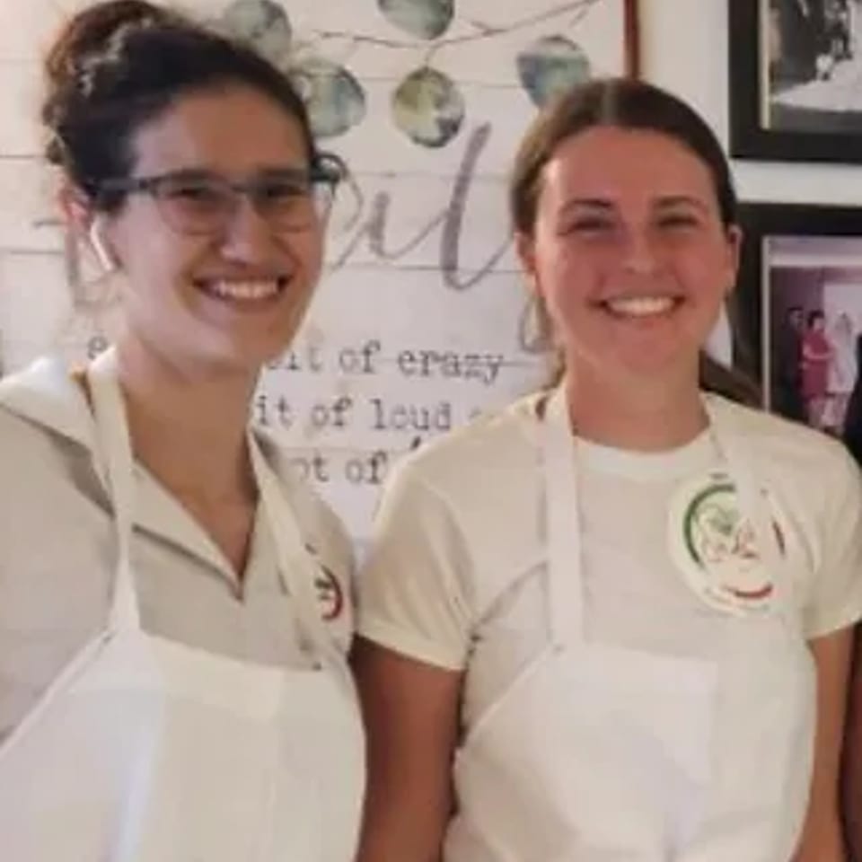 Two smiling women in white aprons, one wearing glasses, standing in front of a decorative wall.