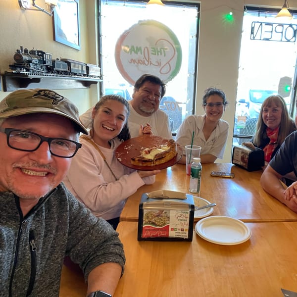 A group of happy people at The Mad Sicilian restaurant, with one person holding a deep-dish pizza.
