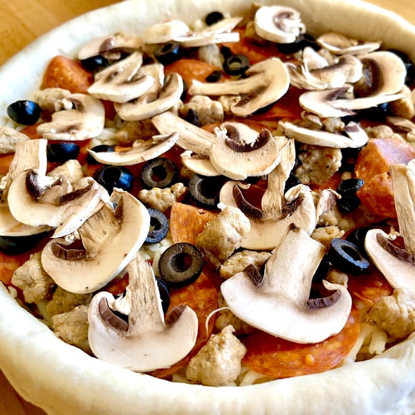 Unbaked deep-dish pizza with pepperoni, sausage, black olives, and mushrooms, ready for the oven.