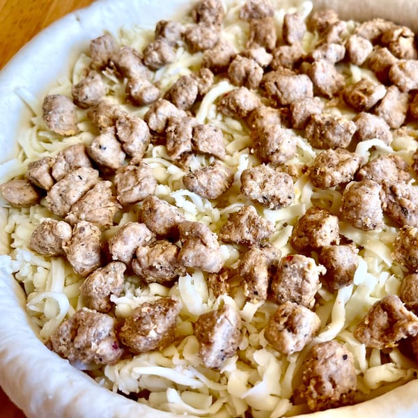 Uncooked pizza with a thick crust, topped with shredded mozzarella cheese and crumbled sausage.