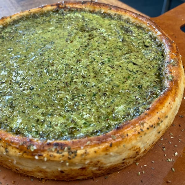 A deep-dish pizza from The Mad Sicilian, topped with a vibrant green pesto sauce and herbs.