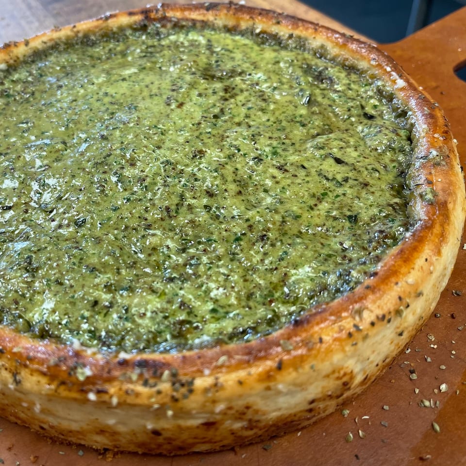 A deep-dish pizza from The Mad Sicilian, topped with a vibrant green pesto sauce and herbs.
