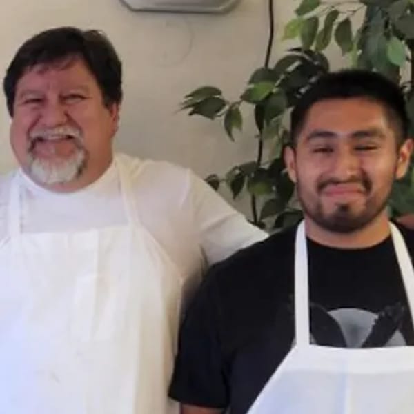 Two smiling men, one older with a beard and one younger, wearing white aprons.