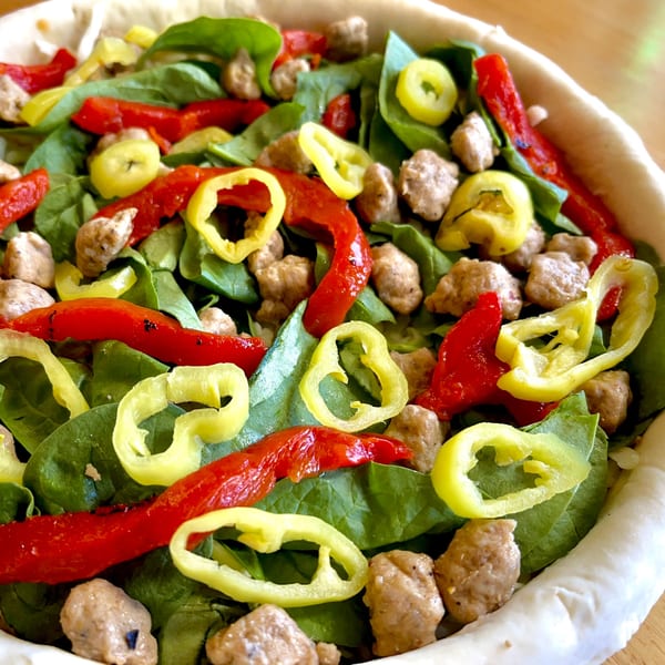 Unbaked deep-dish pizza with spinach, sausage, red peppers, and banana peppers in a crust.