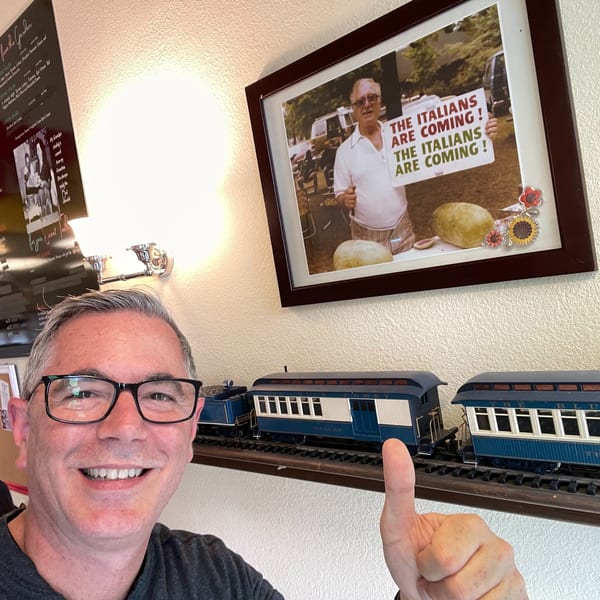 Man smiling and giving a thumbs-up, with a framed Italian-themed photo and model train behind him.